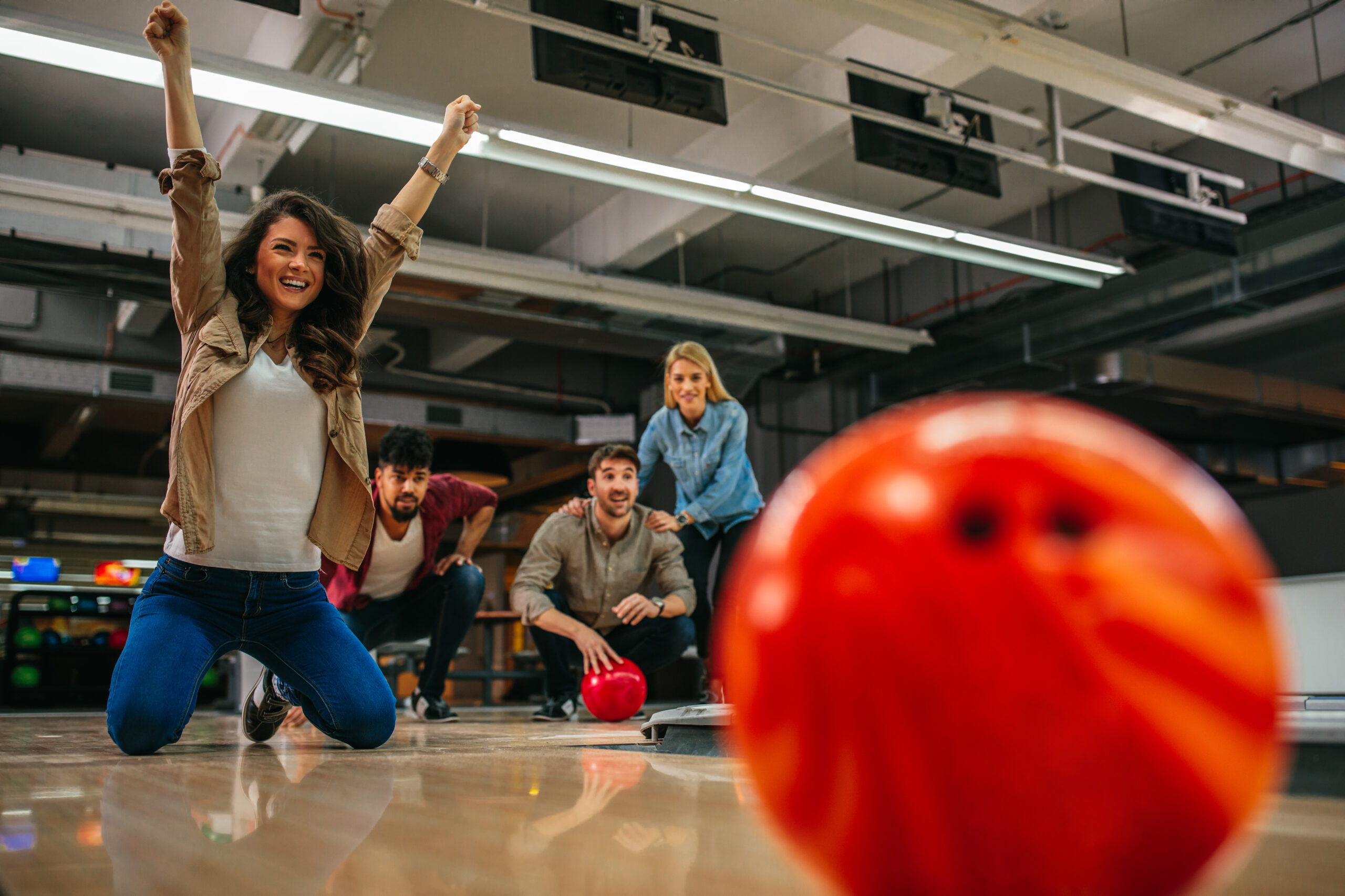 Strike! Bowling-Moment voller Jubel Eine Frau kniet jubelnd auf einer Bowlingbahn, im Hintergrund feuert eine Gruppe Freunde sie an.