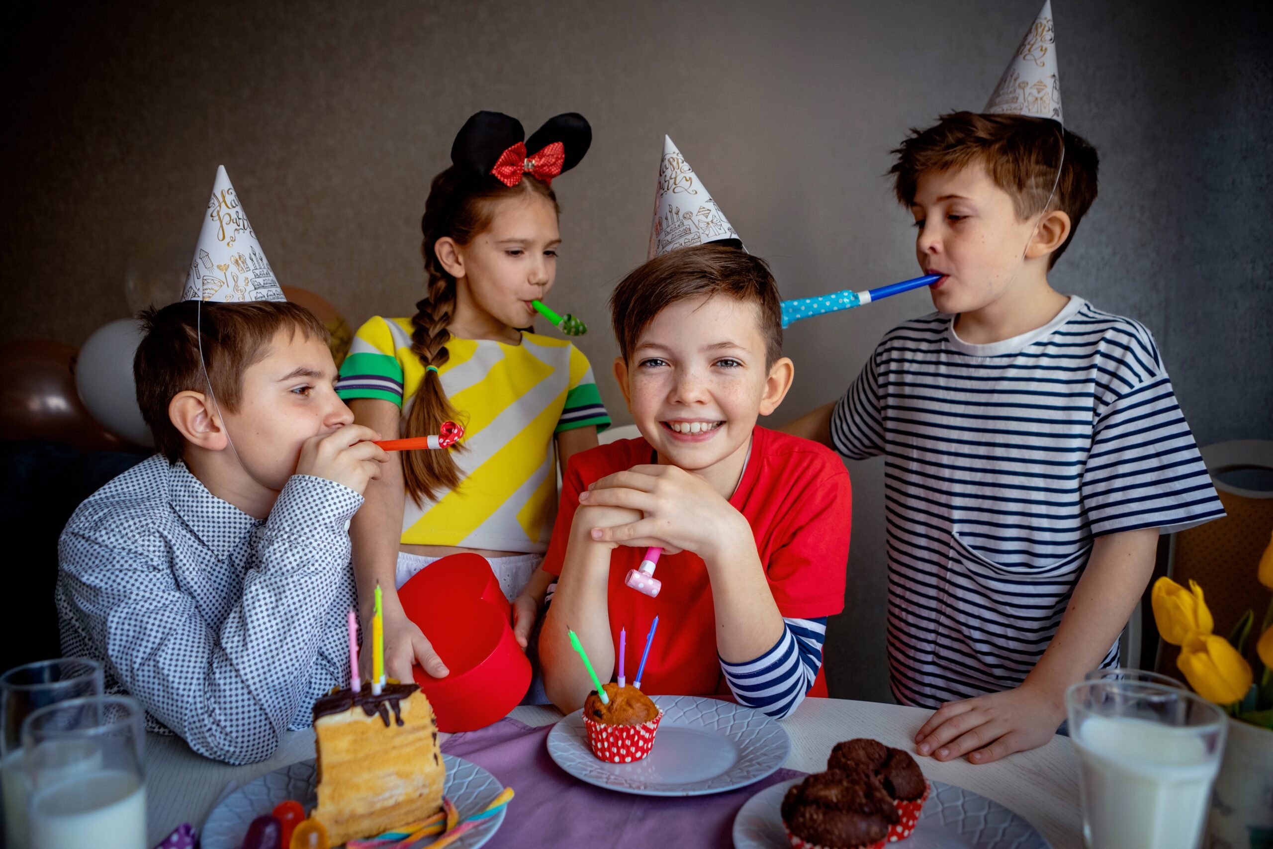 Vier Kinder feiern mit Partyhüten, Kuchen und Lufttröten an einem Geburtstagstisch.
