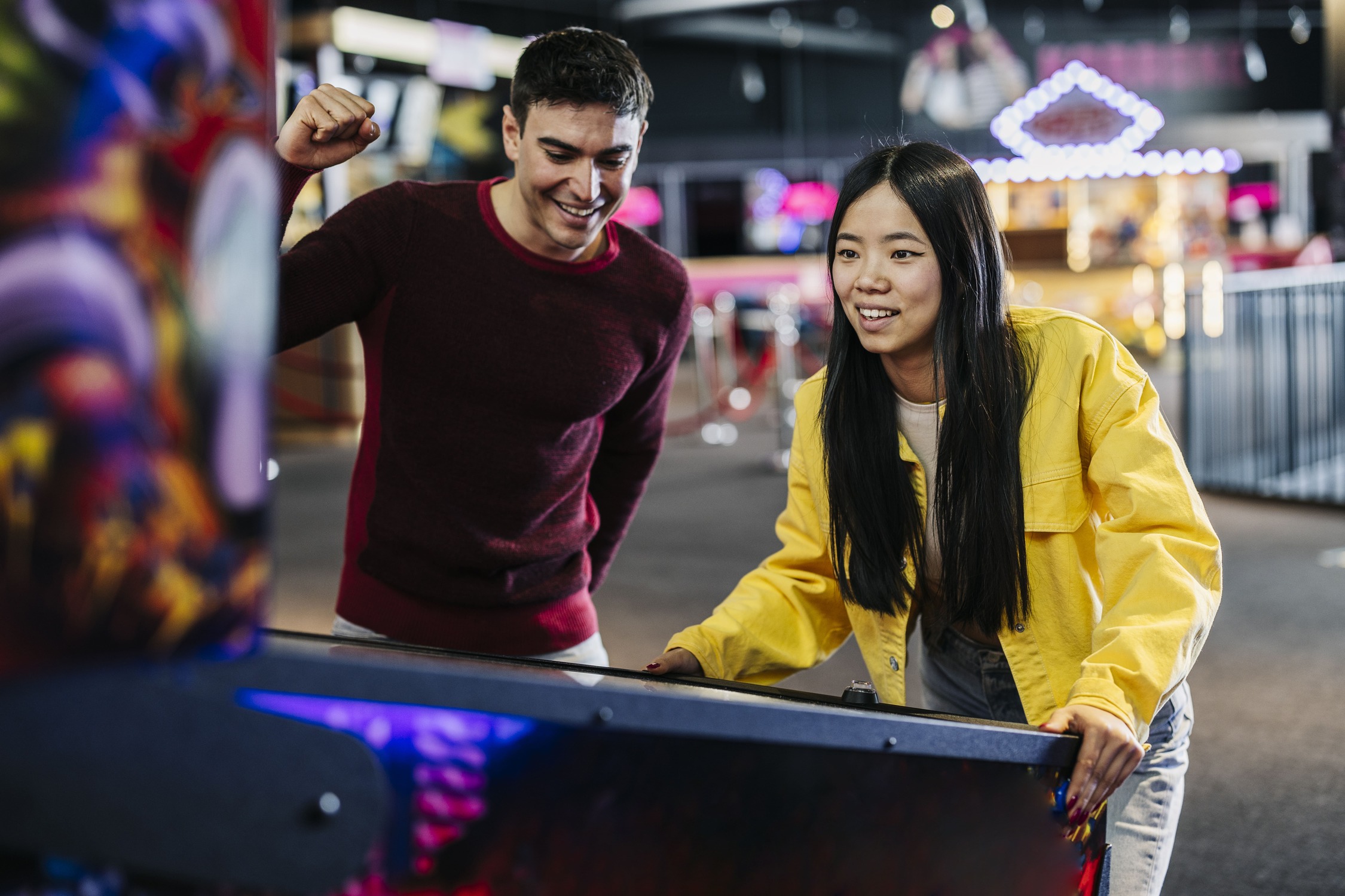 Eine junge Frau spielt konzentriert an einem Flipperautomaten, während ein Mann daneben mitfiebert.