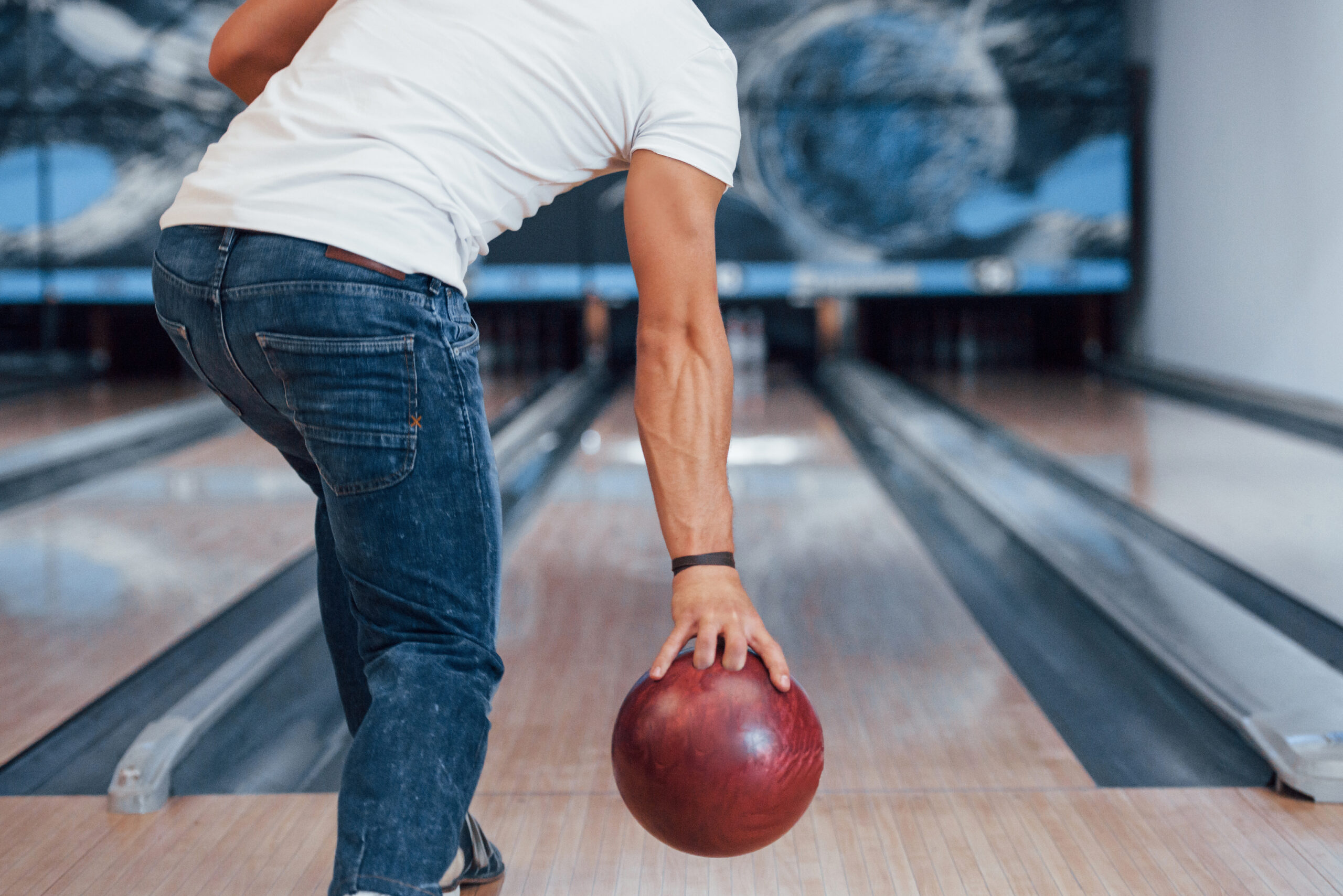 Ein Mann in Jeans und T-Shirt wirft eine rote Bowlingkugel auf einer Bowlingbahn.