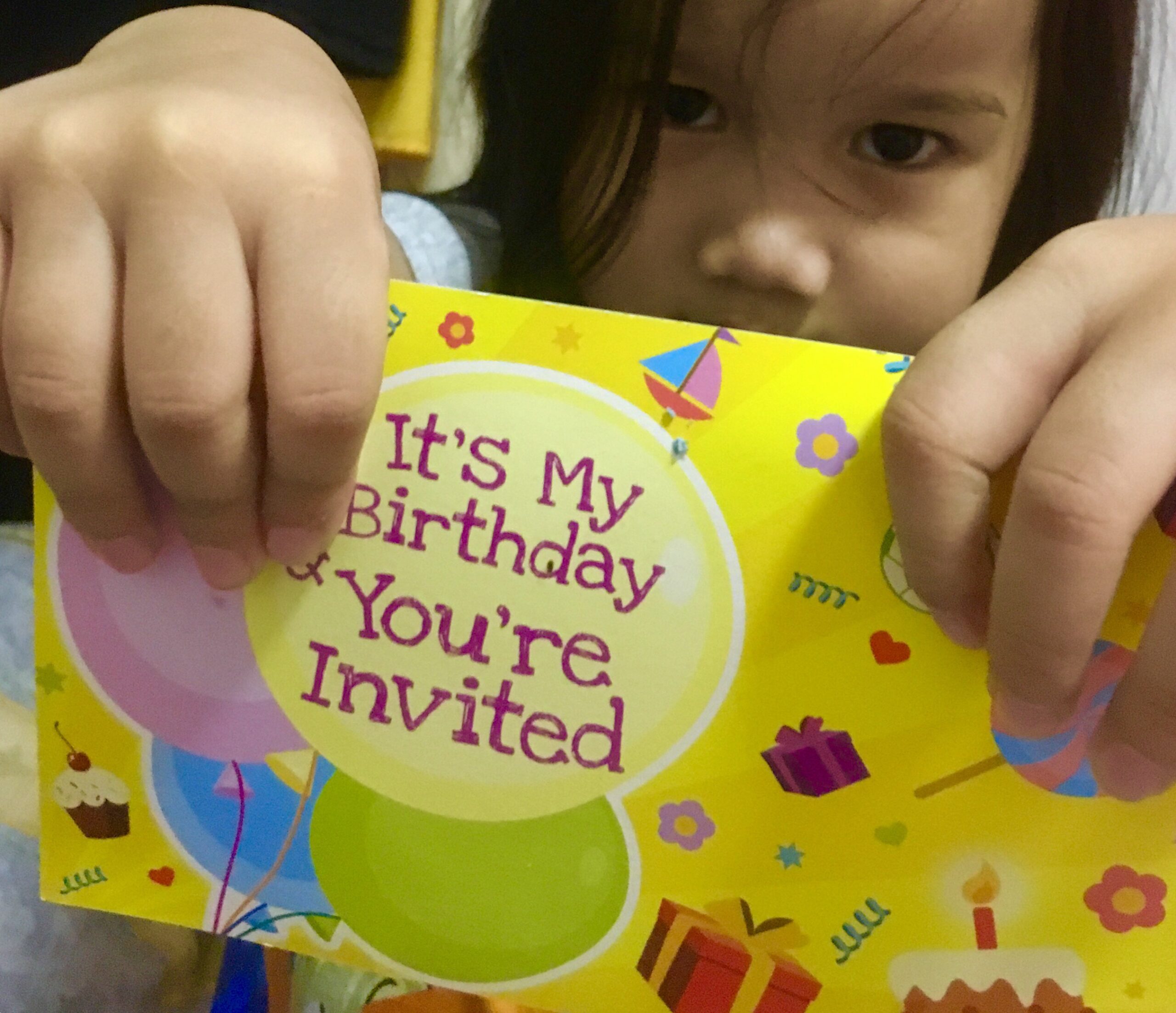 Ein Kind hält eine bunte Einladungskarte mit der Aufschrift „It’s My Birthday & You’re Invited“ in die Kamera.