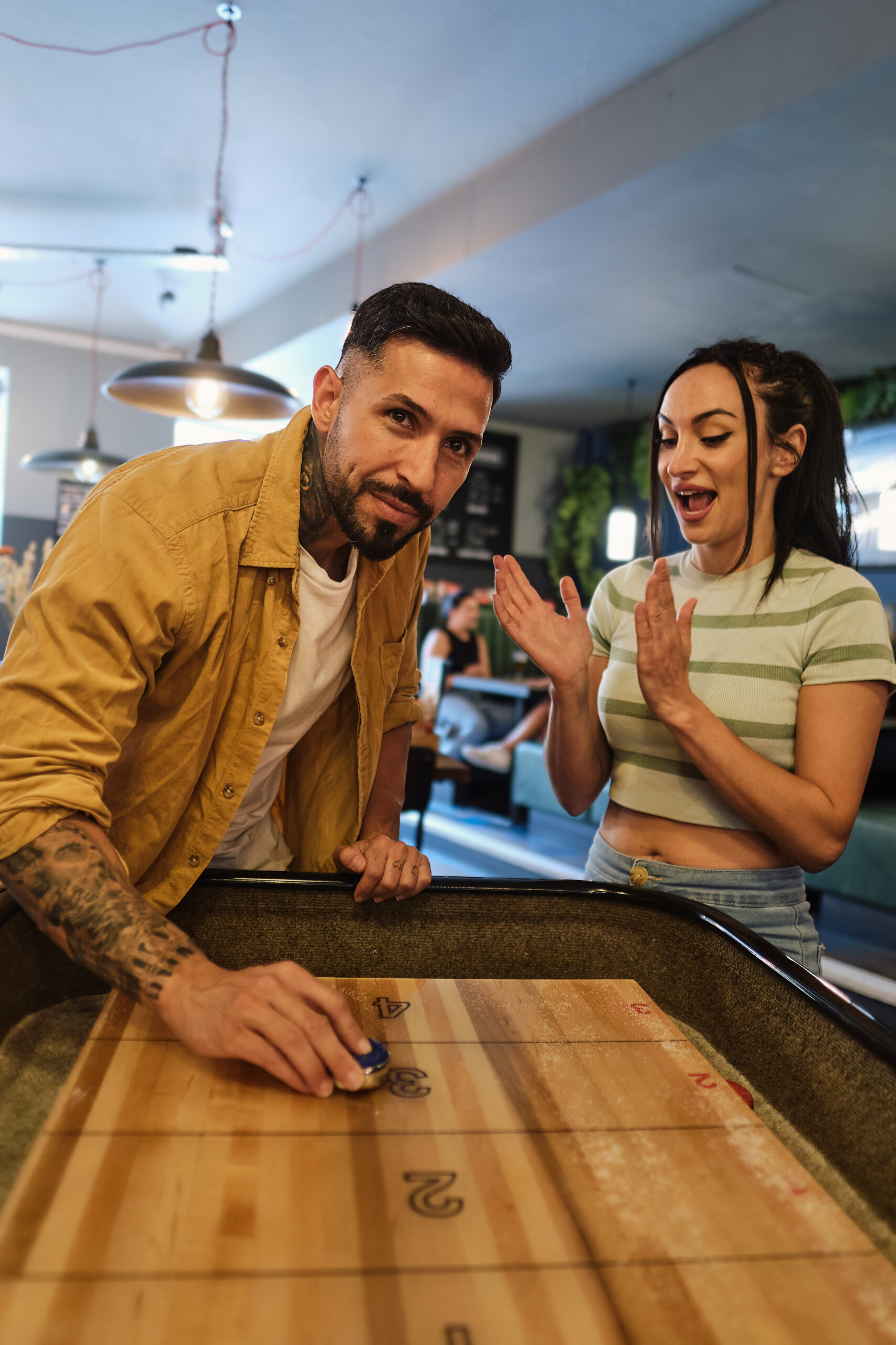Ein Mann schiebt konzentriert eine Shuffleboard-Scheibe, während eine Frau neben ihm applaudiert.